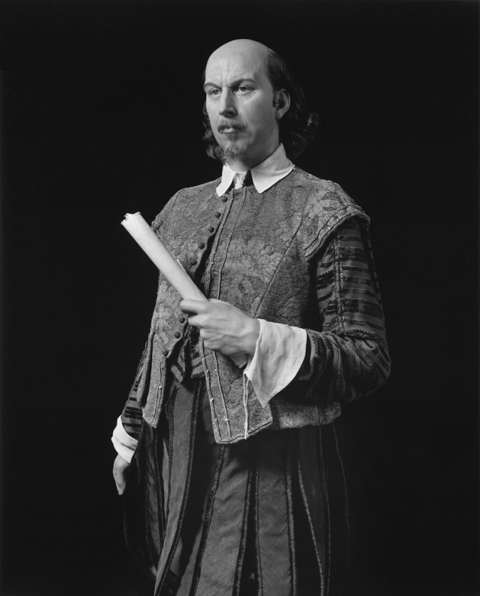 Black-and-white frontal three quarter of a wax figure of a man in a brocaded outfit holding a roll of paper to his torso