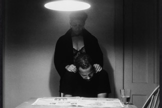 Black and white photograph of an African American couple at a kitchen table, a man seated and reading the paper, a woman standing behind with her hands on his shoulders.