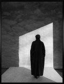 Black and white photograph of a figure in a black coat looking towards a wall of clouds in the sky