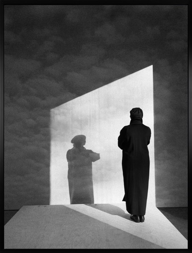Black and white photograph of a figure dressed in black looking at a wall with clouds and their shadow