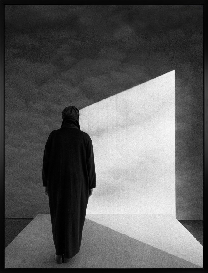 Black and white photograph of a figure dressed in black on wooden platform looking at a wall of clouds