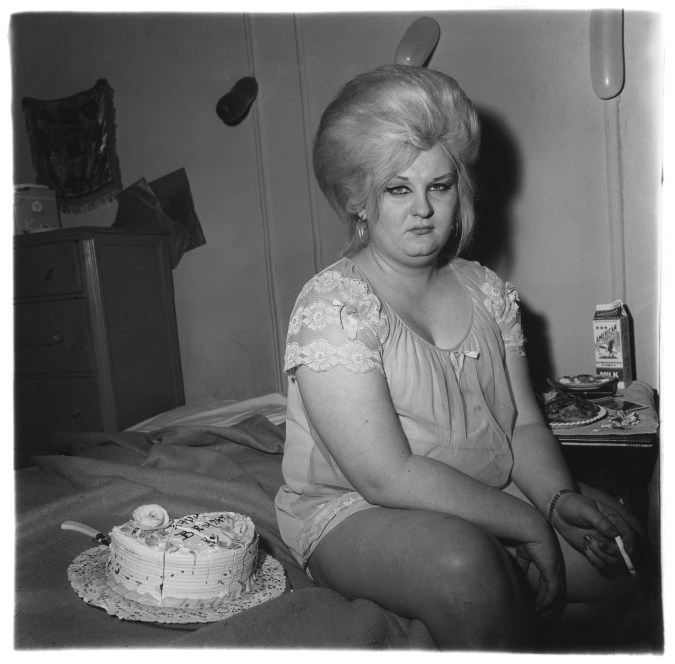A black and white photograph of a feminine figure in a nightgown holding a cigarette while seated on a bed next to a birthday cake