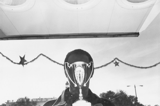 A black and white photograph of the reflection of the artist over a shining trophy in a store window