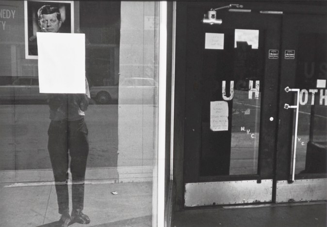 A black and white photograph of a self portrait of the artist reflected in a shop window