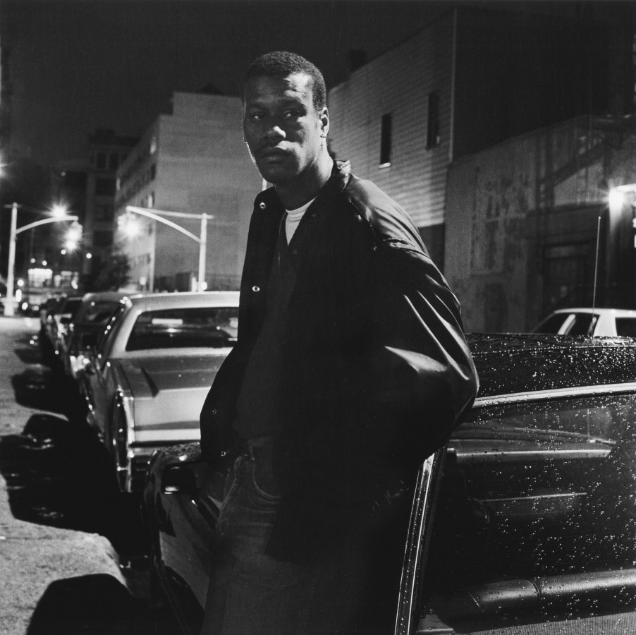 Black and white photograph of a figure leaning against a parked car