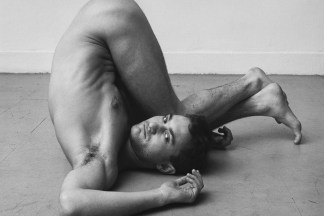 Black-and-white photograph of a contorted nude man with his head and shoulders on the floor and his legs folded back over his torso