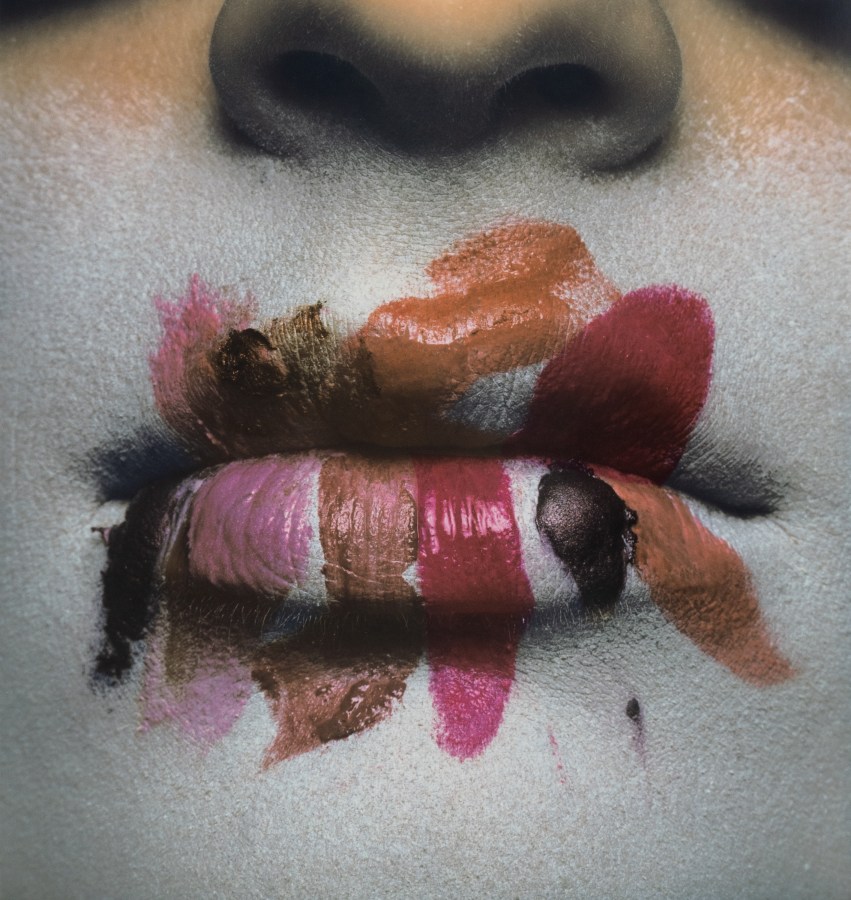 Color photograph of an up close shot of a figures mouth with multiple swatches of lipstick on their lips