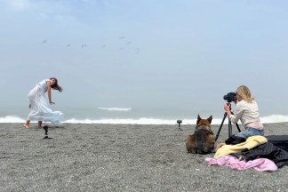 A color image of a photographer shooting pictures of a person wearing a dress on a sandy beach, a dog sits nearby