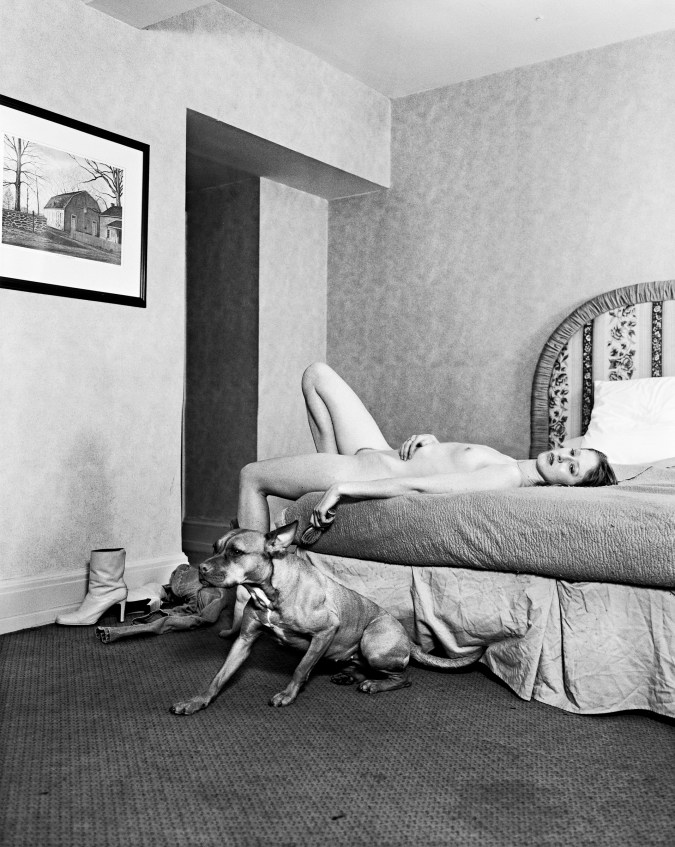 Black-and-white photograph of a nude woman lying on a bed with a dog seated on the floor beside her