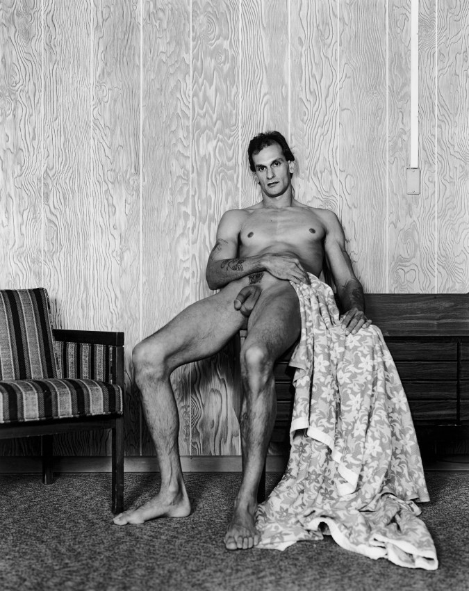Black-and-white photograph of a nude man leaning against a wood paneled wall with a flowered blanket draped by his side
