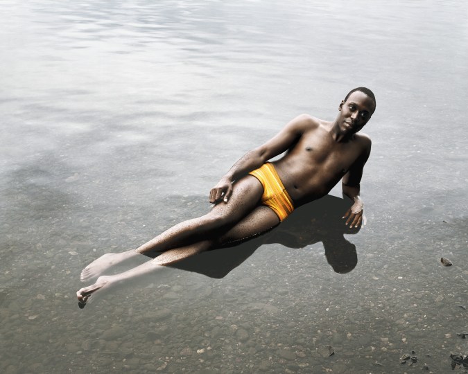 Color photograph of figure laying in shallow water with mustard colored bathing suit