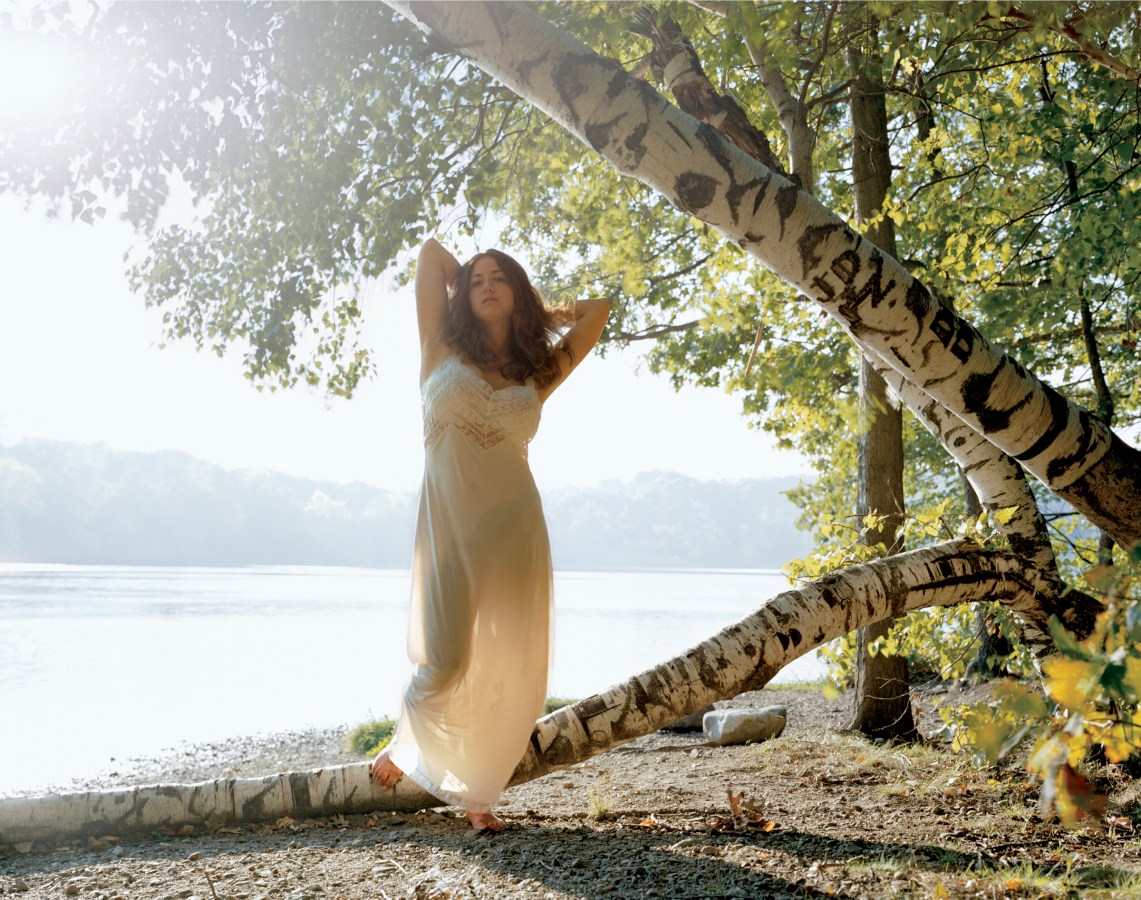 Color photograph of a woman in a long white dress standing on a fallen tree trunk on the shore of a still lake