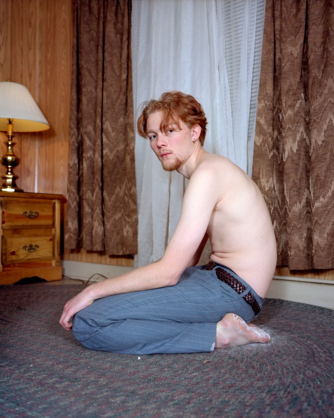 Color photograph of a shirtless young man kneeling on a rug in a wood paneled room