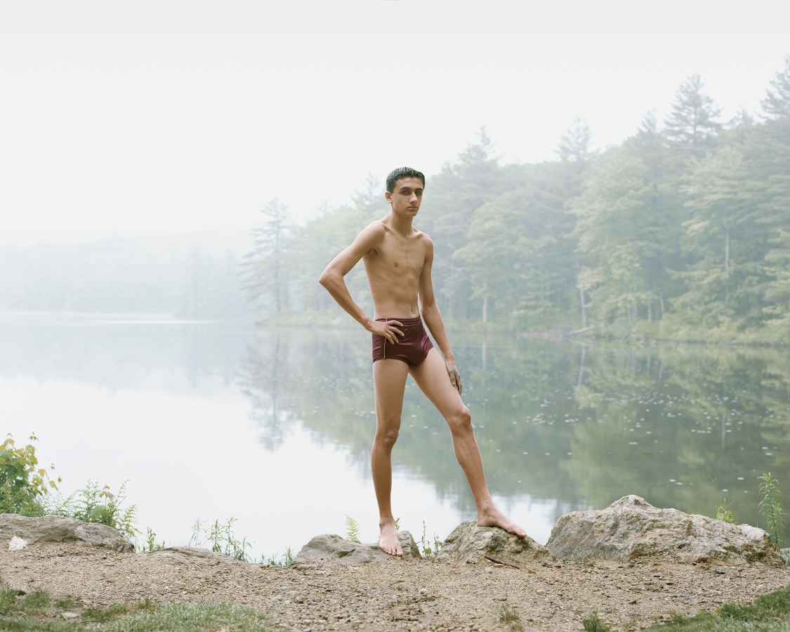 Color photograph of a young man in a bathing suit posed with his hand on his hip on the rocky shore of a misty lake