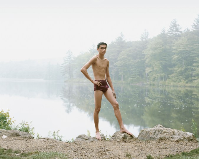 Color photograph of a young man in a bathing suit posed with his hand on his hip on the rocky shore of a misty lake