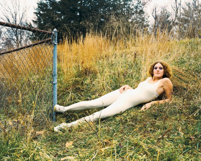 Color photograph of a figure laying a hill of grass with their foot against a metal fence post