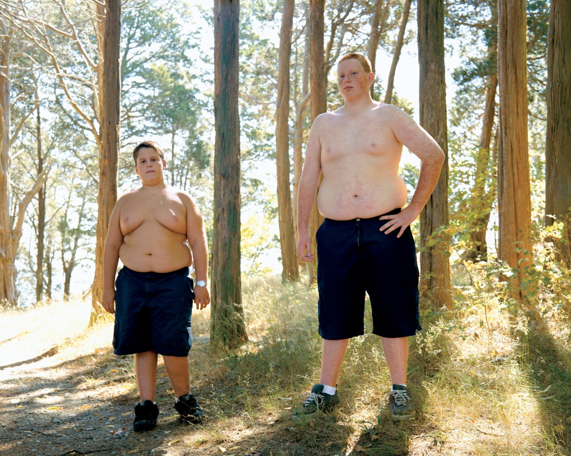 Color photograph of two boys wearing only shorts and sneakers standing in a sunny grove of trees