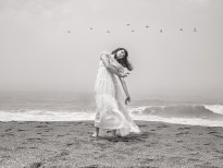 Black and white photograph of a figure in a white dress dancing on the beach as birds fly over head