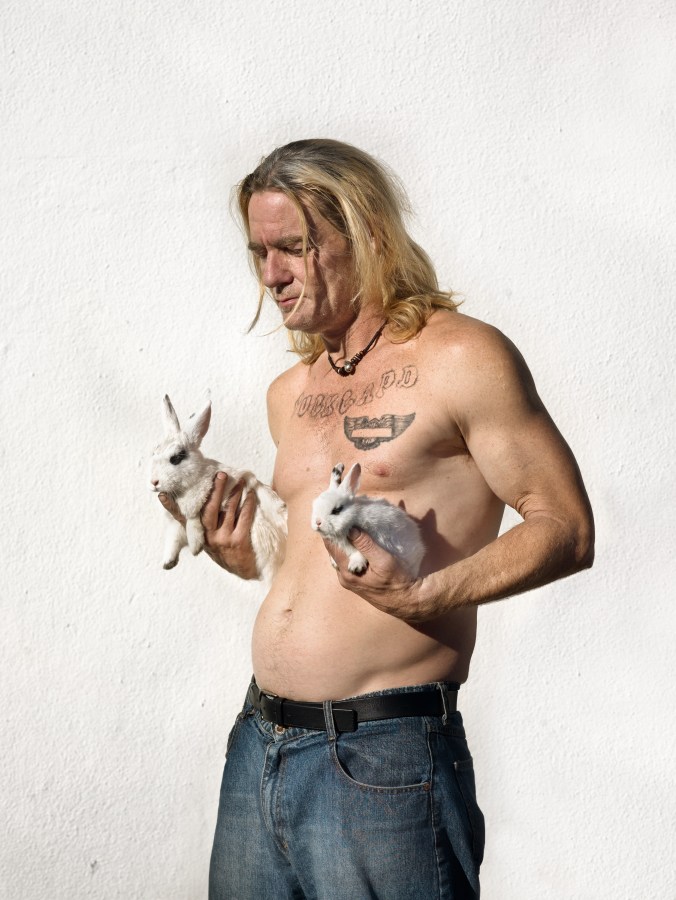 Color photograph of a shirtless figure holding a rabbit in each hand against a textured white wall