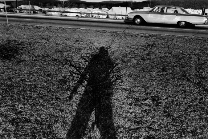 A black and white photograph of the artists shadow with a speeding car in the background.