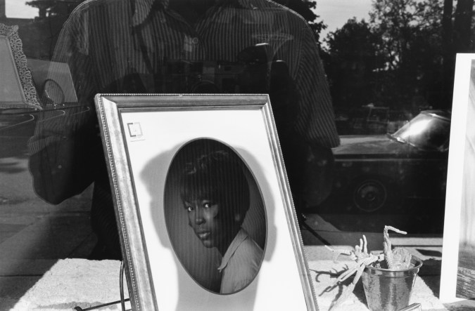 Black and white photograph showing a storefront reflection, with the shadow of the photographer cast onto a framed portrait of a woman.