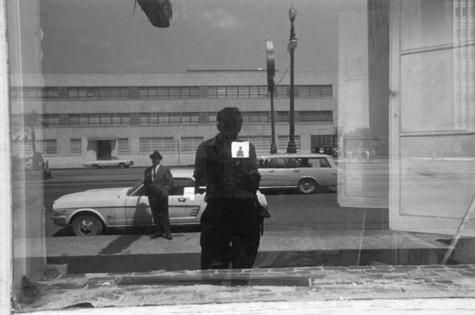 A black and white photograph of the artist reflected in a shop window.