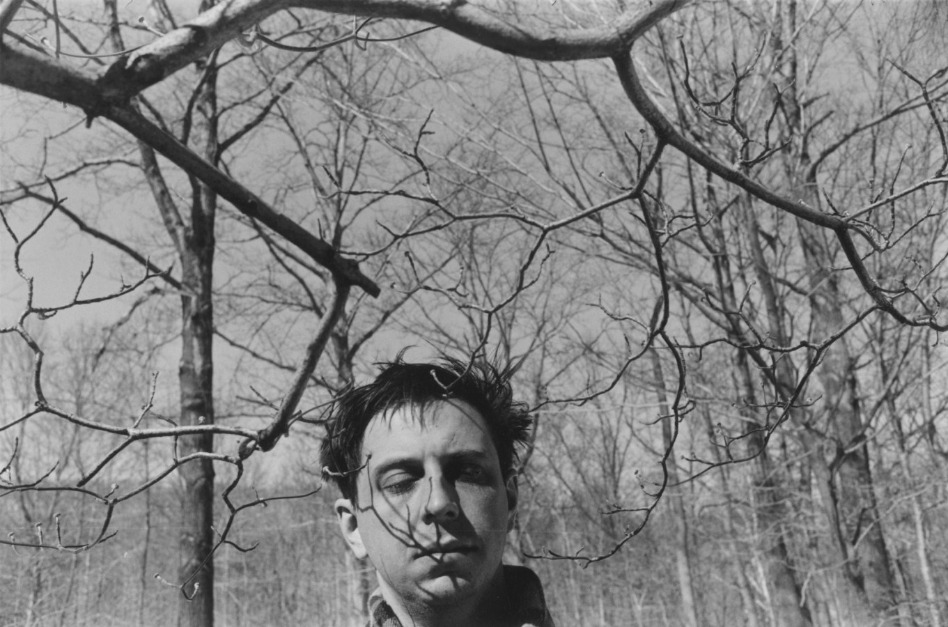 A black and white portrait of the artist with his eyes closed and tree branches in the foreground and background.