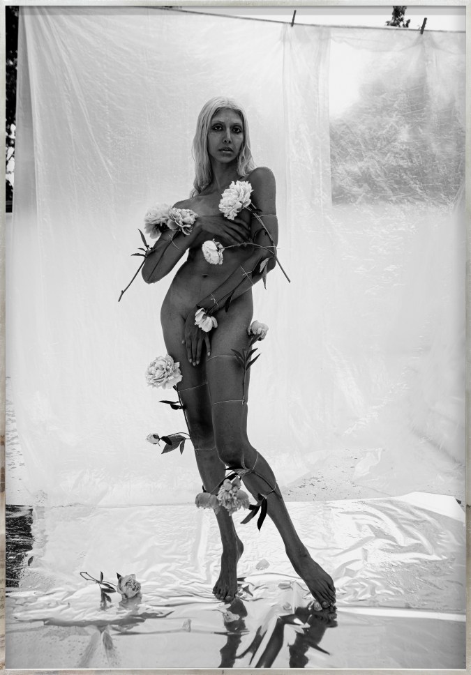 Color image of a black and white photograph of a nude figure covered with flowers and covering their body with their hands framed in distressed metal