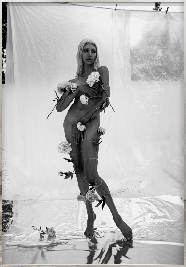 Color image of a black and white photograph of a nude figure covered with flowers and covering their body with their hands framed in distressed metal