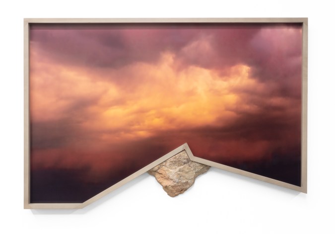 Color photograph of the sky with hues of orange, purple, and red framed in a custom wooden frame with a modified rock puncturing the frame