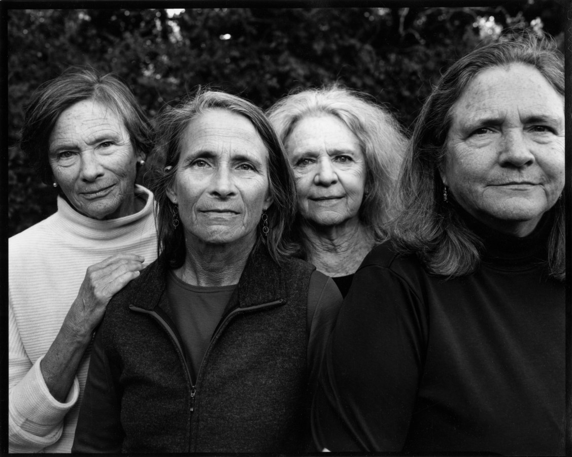 Black and white photograph of four figures in a row looking at the camera with trees behind them