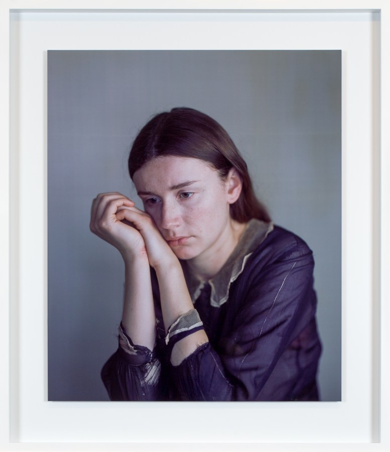 Color photographic portrait of a young woman in a purple shirt with her cheek resting on her clasped hands