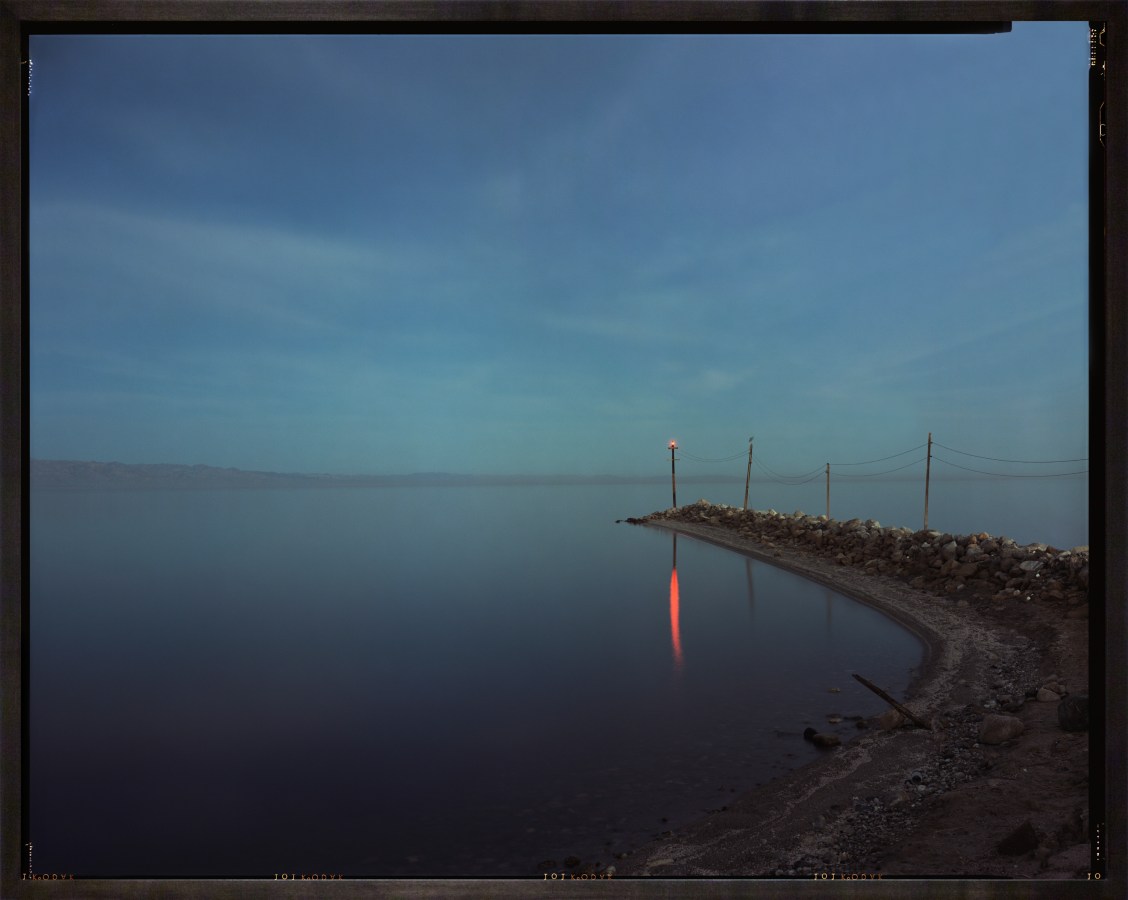 Color photograph of a body of water with red light on a pole reflected in the water framed in a dark wood