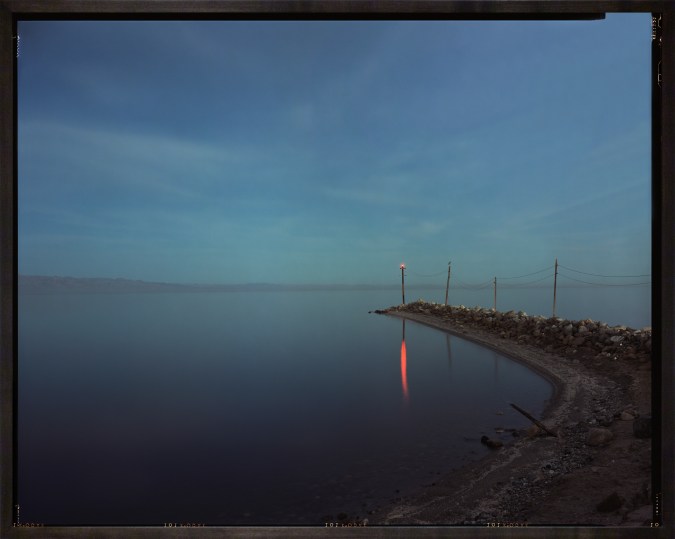 Color photograph of a body of water with red light on a pole reflected in the water framed in a dark wood