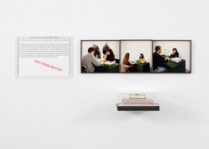 Color image of an artwork including a text panel, a triptych of the artist seated with three different figures, and a stack of paper encased in a plexi box