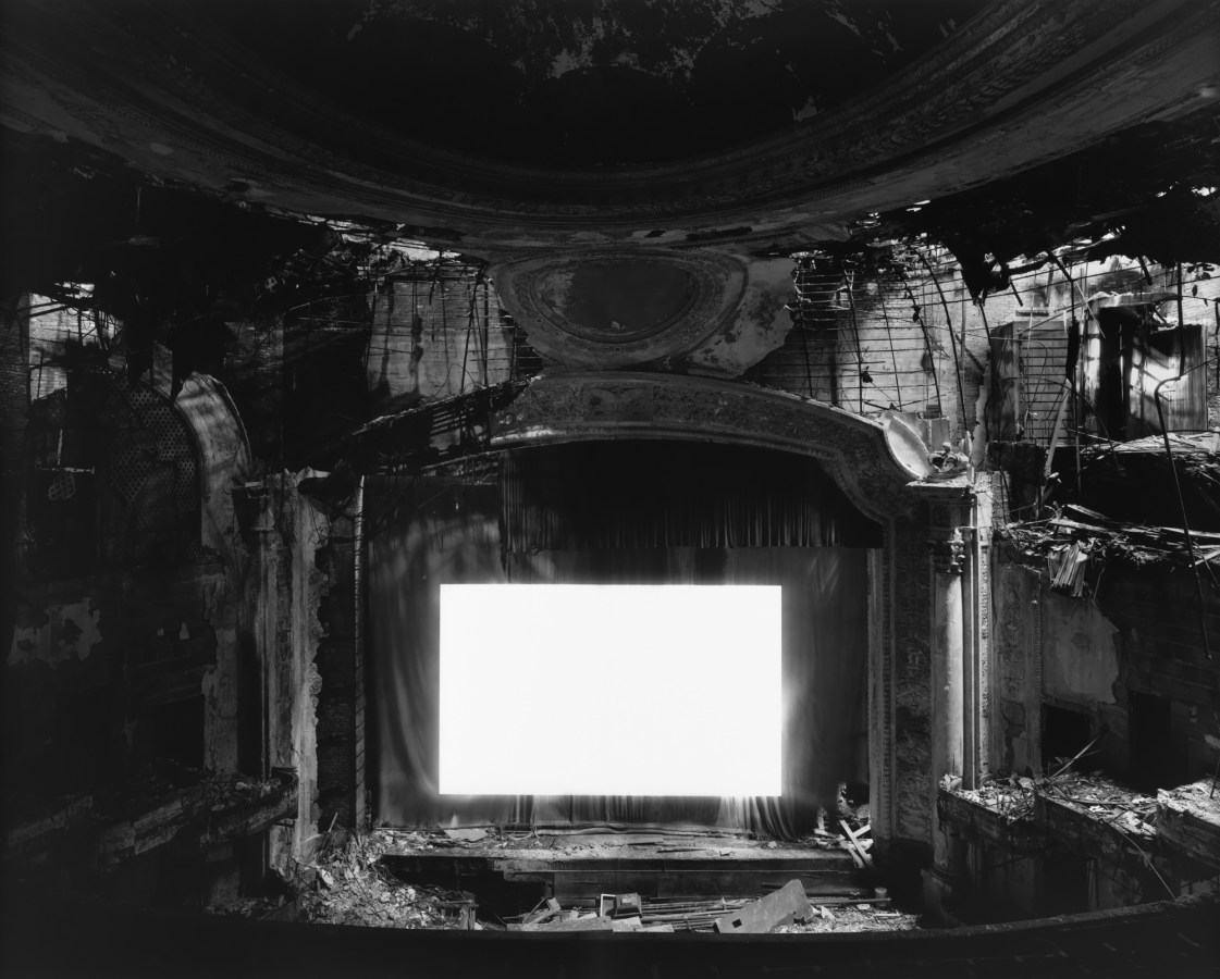Black-and-white photograph of an derelict abandoned theater with a glowing blank white screen