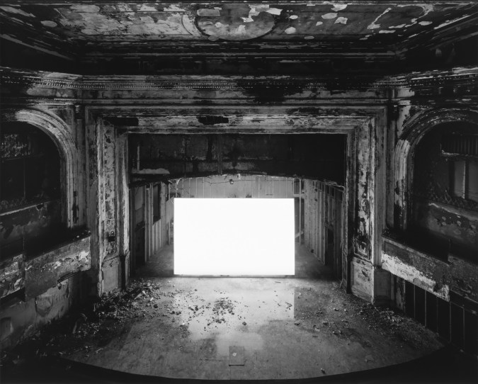 Black-and-white photograph of an empty abandoned theater with a glowing blank white screen
