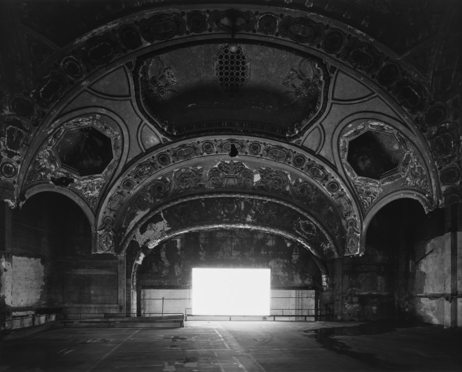 Black-and-white photograph of an empty abandoned theater with a glowing blank white screen