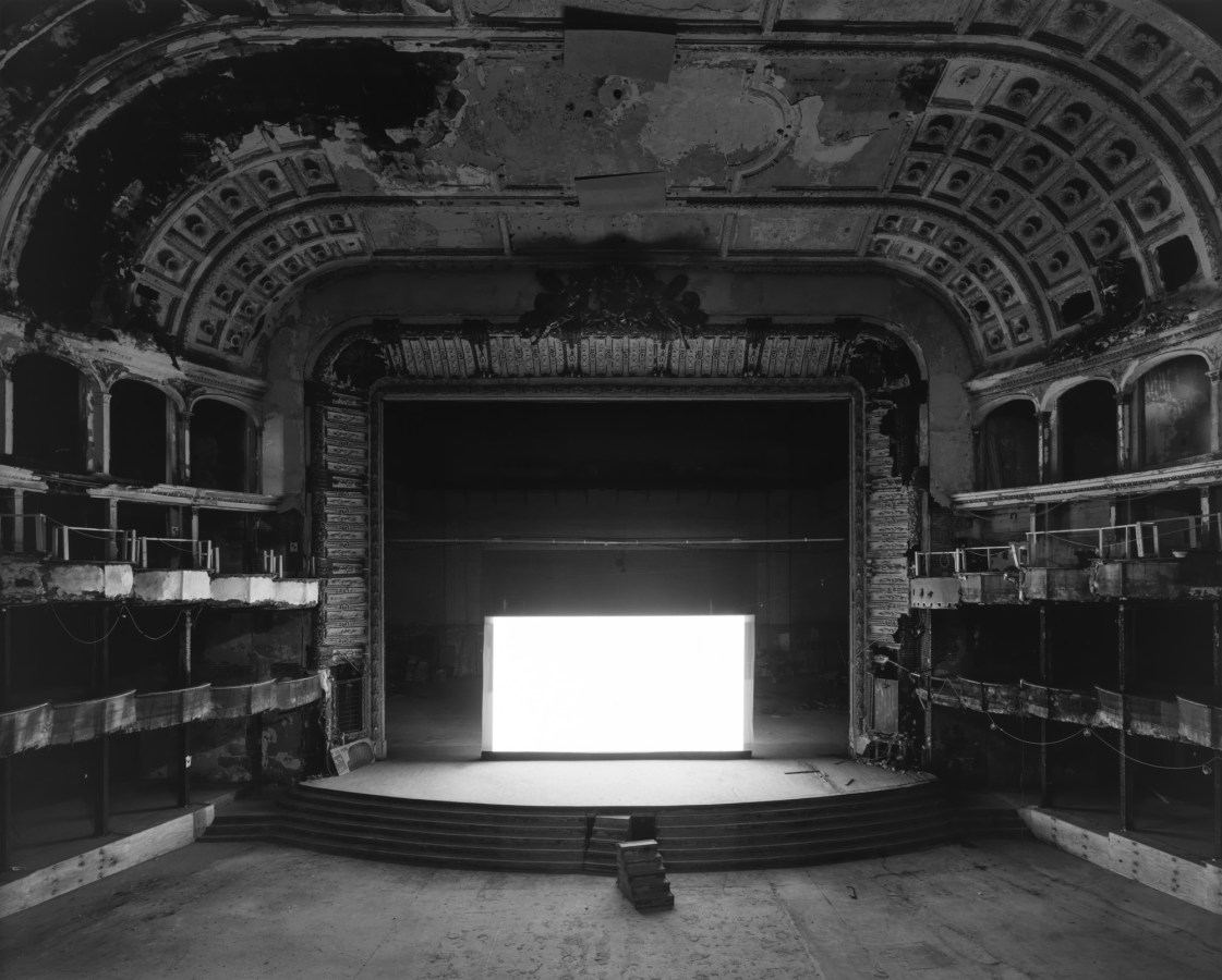 Black-and-white photograph of an empty abandoned theater with a glowing blank white screen