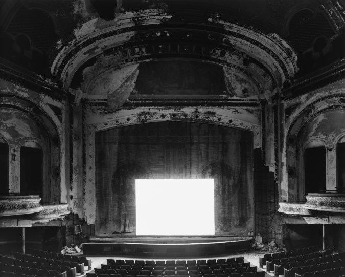 Black-and-white photograph of an empty abandoned theater with a glowing blank white screen